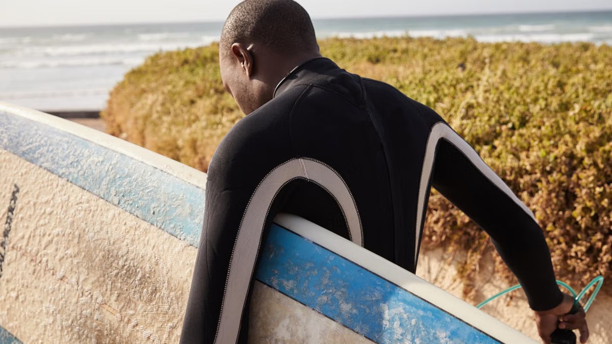 Back view of anonymous ethnic sportsman in diving suit carrying surfboard near shrubs and ocean